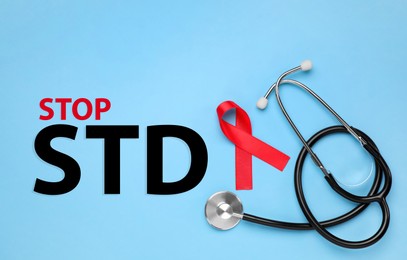 Red awareness ribbon, stethoscope and text Stop STD on light blue background, flat lay