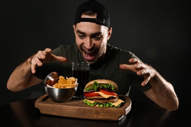 Young hungry man and tasty burger served on wooden board with French fries against black background