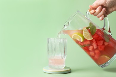 Woman pouring tasty watermelon drink with lime from jug into glass on pale light green background, closeup