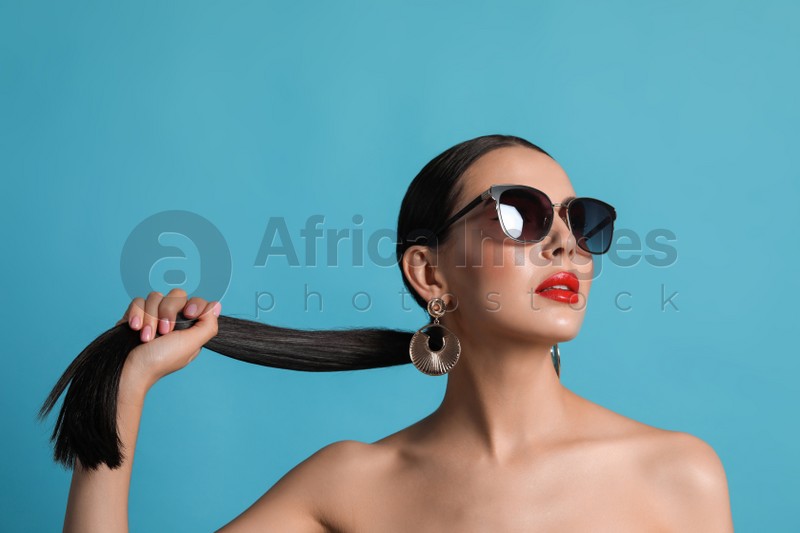 Attractive woman in fashionable sunglasses touching her hair against light blue background