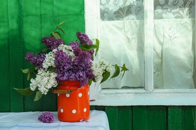 Beautiful lilac flowers in milk can near house outdoors. Space for text
