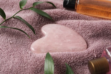 Photo of Rose quartz gua sha tool, green branches and cosmetic products on towel, closeup