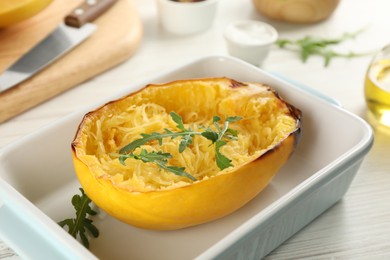 Half of cooked spaghetti squash with arugula in baking dish on white wooden table, closeup