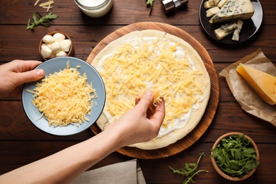Woman adding grated cheese to unbaked pizza on wooden table, top view