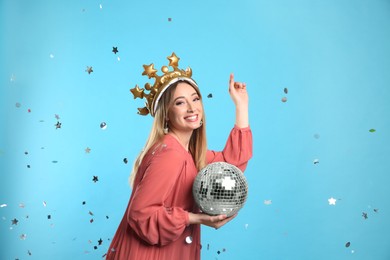 Happy young woman in party crown with disco ball and confetti on light blue background