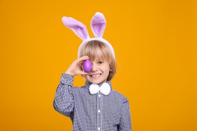 Photo of Happy boy in bunny ears headband holding painted Easter egg on orange background
