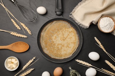 Photo of Frying pan with delicious crepe, spikelets and ingredients on black table, flat lay