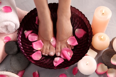 Woman soaking her feet in bowl with water and rose petals on grey floor, top view. Spa treatment