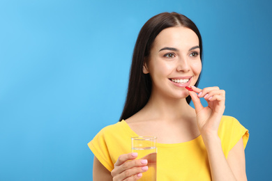Photo of Young woman with glass of water taking vitamin capsule on light blue background. Space for text