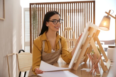 Young woman drawing on easel at table indoors