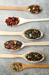 Different kinds of dry herbal tea in wooden spoons on light grey table, flat lay