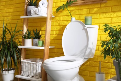 Stylish bathroom with toilet bowl and green plants near yellow brick wall. Interior design