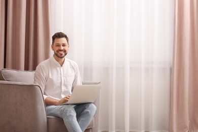 Smiling man holding laptop on armchair near window with beautiful curtains at home. Space for text