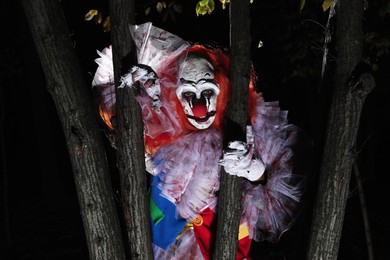 Terrifying clown hiding behind trees outdoors at night. Halloween party costume