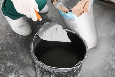 Worker with cement powder and trowel mixing concrete in bucket indoors, closeup