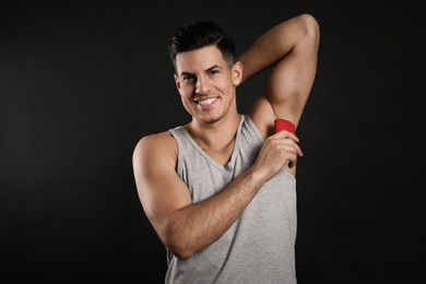 Handsome man applying deodorant to armpit on black background