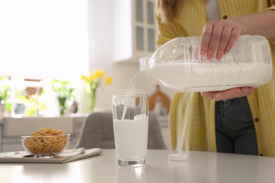 Young woman pouring milk from gallon bottle into glass at white table in kitchen, closeup