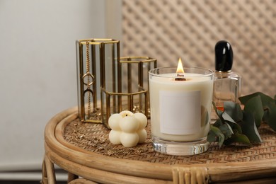 Photo of Burning soy candle, perfume and stylish accessories on wicker table indoors