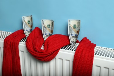 Modern radiator with knitted scarf and money near light blue wall indoors. Energy crisis concept