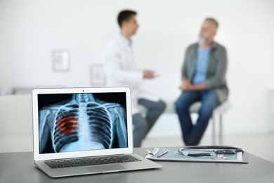 Image of Doctor consulting man in clinic, blurred view. Focus on laptop displaying x-ray of patient with lung cancer