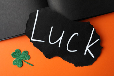 Sheet of paper with word LUCK and clover leaf on orange background, flat lay