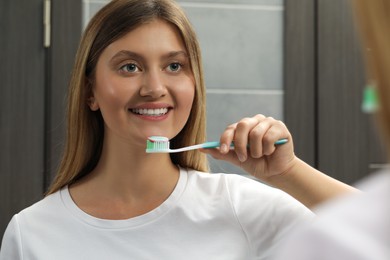 Young woman holding brush with toothpaste near mirror in bathroom