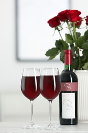 Bottle, glasses of red wine and vase with roses on table in room. Romantic date
