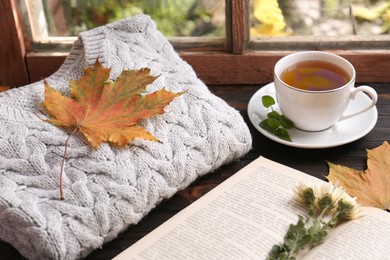 Photo of Cup of aromatic tea, soft sweater and book on wooden windowsill indoors. Autumn atmosphere