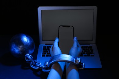 Photo of Woman shackled with ball and chain holding smartphone near laptop on dark background, closeup. Internet addiction