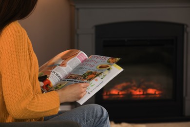 Young woman reading magazine near fireplace at home, closeup. Space for text