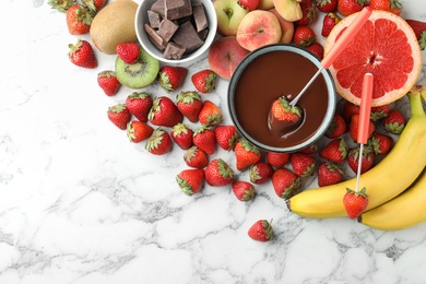 Fondue fork with strawberry in bowl of melted chocolate surrounded by other fruits on white marble table, flat lay