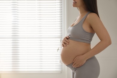 Pregnant young woman touching belly near window indoors, closeup. Space for text