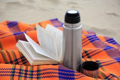 Photo of Metallic thermos with hot drink, open book and plaid on sandy beach, closeup