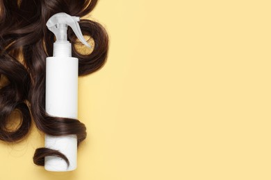 Photo of Spray bottle with thermal protection wrapped in lock of brown hair on yellow background, flat lay. Space for text