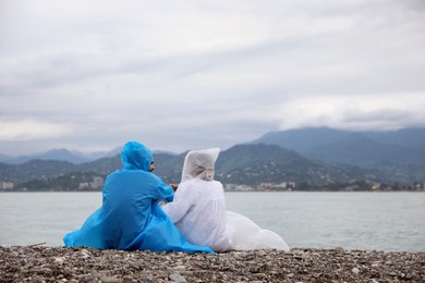Photo of Young couple in raincoats enjoying time together under rain on beach, back view. Space for text