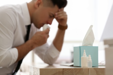 Ill businessman in office, focus on table with box of paper tissues, nasal spray and drops