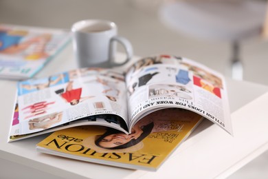 Photo of Fashion magazines and cup of hot drink on white table indoors, closeup