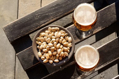 Photo of Glasses of cold beer and pistachios on wooden bench, flat lay
