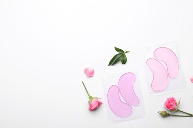 Packages with under eye patches and rose flowers on white background, flat lay. Space for text