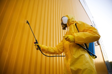 Person in hazmat suit disinfecting building wall with sprayer outdoors. Surface treatment during coronavirus pandemic