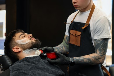 Professional hairdresser applying shaving foam onto client's beard in barbershop
