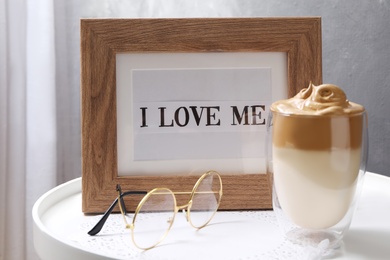 Frame with phrase I Love Me near eyeglasses and cup of coffee drink on table indoors