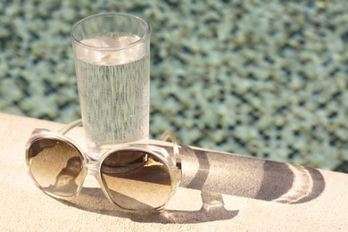 Photo of Stylish sunglasses and glass of water near outdoor swimming pool on sunny day, space for text
