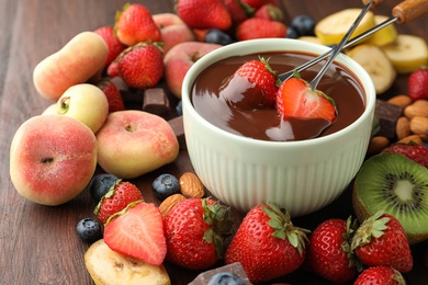 Fondue forks with strawberries in bowl of melted chocolate surrounded by other fruits on wooden table
