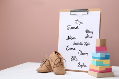 Clipboard with written different baby names, child's shoes and wooden toy on white wooden table