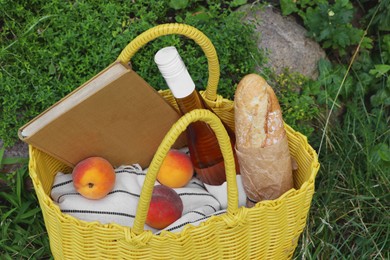 Photo of Yellow wicker bag with book, peaches, baguette and wine on green grass outdoors