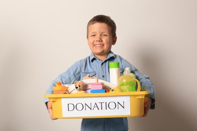 Cute little boy holding donation box with goods and toys against light background