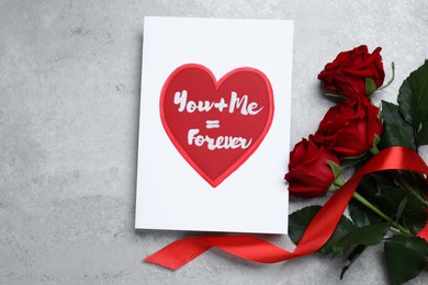 Card with love message near red roses on grey table, flat lay