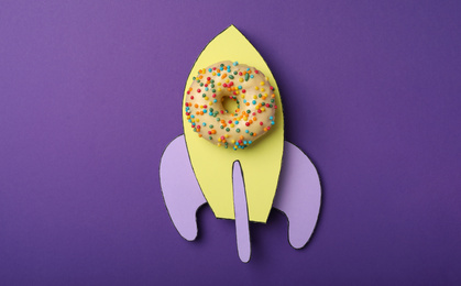 Rocket made with donut and paper on purple background, top view