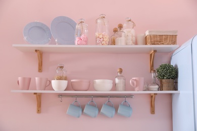 Shelves with dishware and products on pink wall in kitchen